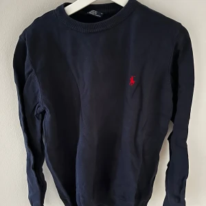 Mörkblå tröja från Ralph Lauren - Säljer en stilren mörkblå tröja från Ralph Lauren med det klassiska röda logotypbroderiet på bröstet. Tröjan har långa ärmar och en rund halsringning. Perfekt för en avslappnad men ändå snygg look.