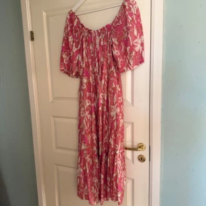 Rosa blommig klänning - Charmig rosa klänning med blommigt mönster och puffiga kortärmar. Den har en luftig passform och en offshoulder-design som ger en romantisk känsla. Perfekt för sommarens varma dagar.