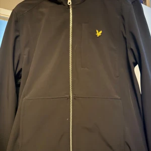 Svart jacka från Lyle & Scott - Snygg svart jacka från Lyle & Scott med dragkedja och huva. Jackan har en broderad logotyp på bröstet och är perfekt för en stilren look. Köpt föra året för 1400 använd en gång inga hål eller defekter så ny skick 