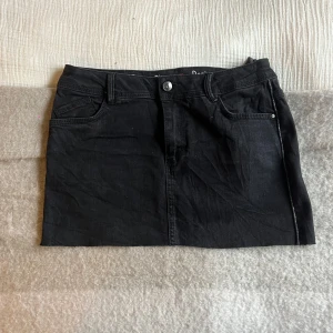 Svart jeanskjol - Snygg svart jeanskjol med klassisk femficksdesign och knappstängning fram. Perfekt för en avslappnad stil. Kjolen har en kort längd och är tillverkad i ett robust denimtyg.