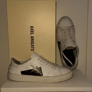 Vita sneakers från Axel Arigato - Snygga vita sneakers från Axel Arigato med svart fågelbroderi på sidan. Skorna har klassisk snörning och en stilren design som passar perfekt till vardags. De är tillverkade i skinn och har en bekväm passform.Skick 7/10. Har använt dem och de sjukt stilrena och unika. Säljer pga av nya skor. Hör av vid frågor eller pris diskussion