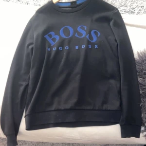 Svart sweatshirt från Hugo Boss - Snygg svart sweatshirt från Hugo Boss med blått tryck på bröstet. Tröjan har långa ärmar och en klassisk rund halsringning. Perfekt för en stilren och avslappnad look. Hör av er vid funderingar eller frågor pris kan diskuteras vid snabb affär tröjan har inga hål finns inget tecken på användning:)