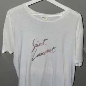 Vit t-shirt från Saint Laurent - Säljer en stilren vit t-shirt från Saint Laurent med deras ikoniska logga i en subtil färgskiftning på bröstet. T-shirten har en klassisk rund hals och korta ärmar. Perfekt för en avslappnad och trendig look.