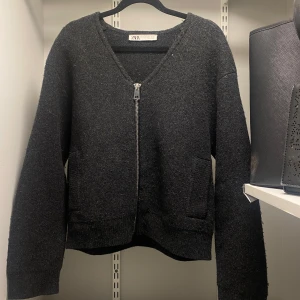Mörkgrå kofta från Zara - Säljer en stilren mörkgrå kofta från Zara med dragkedja framtill. Koftan har en v-ringad design och långa ärmar, perfekt för kyligare dagar. Den är tillverkad i ett mjukt material som ger en bekväm passform.