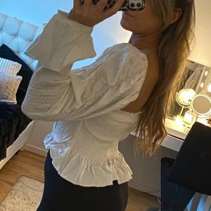 Vit blus med volangdetaljer - Säljer en söt vit blus med volangdetaljer och långa ärmar. Blusen har en smickrande passform med elastiska partier och en lätt rynkad effekt. Perfekt för en stilren look.