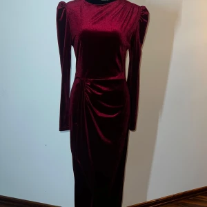 Vinröd sammetsklänning - Elegant vinröd klänning i sammet med långa ärmar och puffiga axlar. Klänningen har en smickrande drapering i midjan och en diskret dragkedja i ryggen. Perfekt för en glamorös kväll.