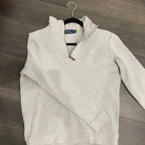 Grå half zip från Ralph Lauren - Säljer en stilren grå tröja från Ralph Lauren med halv dragkedja och broderad logga på bröstet. Perfekt för en avslappnad och snygg look. Tröjan har långa ärmar och är tillverkad i ett mjukt material.