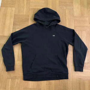 Svart hoodie från Under Armour - Säljer en svart hoodie från Under Armour i storlek M. Den har en klassisk design med en liten logga på bröstet och en praktisk magficka. Perfekt för en avslappnad stil.