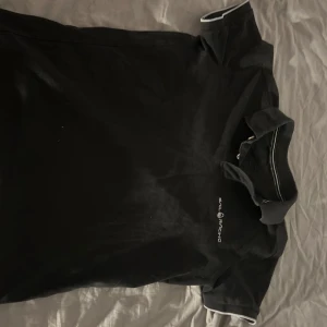 Svart pikétröja från Sail Racing - Snygg svart pikétröja från Sail  Racing med vita detaljer på krage och ärmslut. Perfekt för en stilig look. Kortärmad design med klassisk pikékrage.