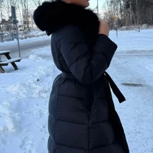 Svart dunjacka med pälsdetalj - Snygg svart dunjacka med en lyxig pälsdetalj på huvan. Jackan har en figursydd passform med ett bälte i midjan för extra stil. Perfekt för kalla vinterdagar.