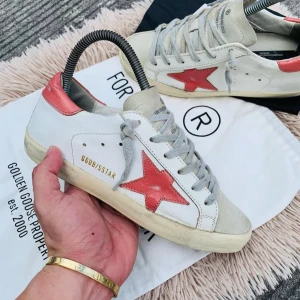 Golden Goose sneakers med röd stjärna - Snygga Golden Goose sneakers i vitt med en röd stjärna på sidan. Skorna har grå snörning och en röd detalj vid hälen. Perfekta för en trendig och avslappnad stil.
