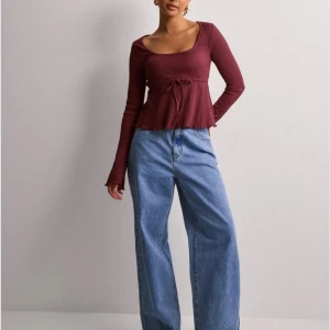 Vinröd ribbad topp med knyt - Snygg vinröd topp med ribbad struktur och knytband i midjan. Toppen har långa ärmar och en rund halsringning. Perfekt för en stilren look. Första bilden är en mer rättvis färg på tröjan än bild 2