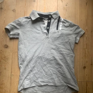 Grå pikétröja från Copperfield - Säljer en stilren grå pikétröja från Copperfield. Tröjan har korta ärmar och en klassisk krage med knappar. Den är tillverkad i bomull och elastan för en bekväm passform. Detaljer som broderad logga på ärmen och randiga accenter vid knapparna ger en snygg touch.