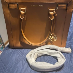 Brun handväska från Michael Kors - Snygg brun handväska från Michael Kors i skinn med guldfärgade detaljer och logotyp. Väskan har dubbla handtag och en praktisk dragkedja upptill. Insidan är fodrad med Michael Kors-mönster och har flera fack för enkel organisering. Bred 30, längd 50