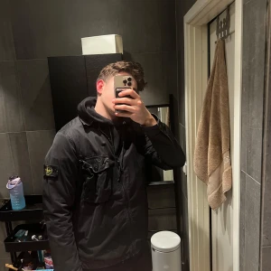 Svart overshirt från Stone Island - Säljer en svart overshirt från Stone Island med dragkedja och broderad logotyp på ärmen. Jackan har en klassisk krage och en ficka på bröstet. Perfekt för en stilren look nu till våren.