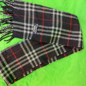 Rutig Halsduk från Burberry - Snygg rutig halsduk från Burberry i 100% lammull. Den har en klassisk design med svarta, grå och röda rutor. Perfekt för att hålla dig varm och stilren under kyliga dagar. Tillverkad i England. Äkta