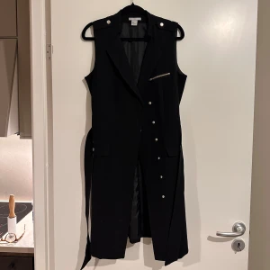 Svart klänning med knappar  - Snygg svart klänning med tryckknappar framtill. Klänningen har en stilren design med ett bälte i midjan och dekorativa knappar på axlarna. Perfekt för att ge en extra touch till din outfit. Aldrig använd, storleken är M och har inga riktiga fickor. 