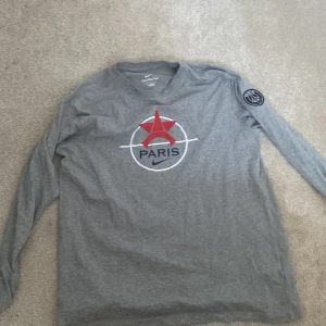 Grå långärmad Nike tröja med Paris-motiv - Säljer en grå långärmad tröja från Nike med ett coolt Paris-motiv i rött och blått på framsidan. Tröjan har en rund hals och ett Paris Saint-Germain-märke på ärmen. Perfekt för fotbollsfans!