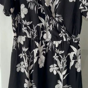 Svart blommig klänning från Only Carmakoma - Snygg svart klänning från Only Carmakoma med ett vackert vitt blommönster. Klänningen har en omlott-design och korta ärmar, perfekt för en stilren look. Den elastiska midjan ger en bekväm passform. Perfekt för att känna sig både bekväm och chic.