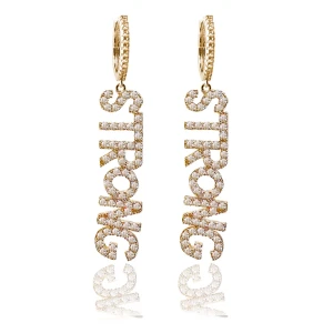 ioaku örghängen - Snygga guldiga örhängen med texten STRONG från ioaku. Perfekta för att göra ett statement och ge en extra touch till din outfit. De hängande bokstäverna är täckta med små gnistrande stenar som fångar ljuset vackert.