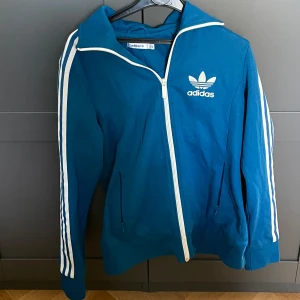 Blå träningsjacka från Adidas - Säljer en blå träningsjacka från Adidas med vita ränder längs ärmarna och dragkedja framtill. Jackan har en klassisk Adidas-logga på bröstet och långa ärmar. 