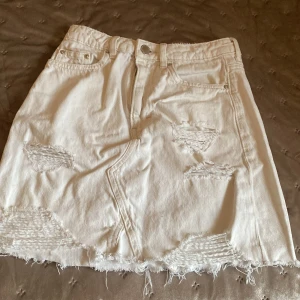 Vit jeanskjol från H&M Divided - Säljer en cool vit jeanskjol från H&M Divided i storlek 34. Kjolen har slitna detaljer och en rå kant nertill som ger en trendig look. Perfekt för att skapa en avslappnad stil. Den har knapp och dragkedja framtill. den är inte sönder. Det är bara design. Sitter som en smäck och är perfekt nu när det börjar bli sommar. 