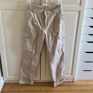 Beige cargobyxor från Nelly  - Snygga beige cargobyxor med flera praktiska fickor och knappdetaljer. Byxorna har en avslappnad passform och är perfekta för en casual look. Tillverkade i ett slitstarkt material som ger en bekväm känsla hela dagen. Storlek 36. Från Nelly.