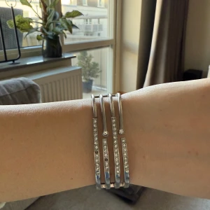 Silverfärgat armband med strassdetaljer - Elegant silverfärgat armband med flera rader av gnistrande strass. Armbandet har en modern design med en stilren låsning på baksidan. Perfekt för att lägga till lite glamour till din outfit.
