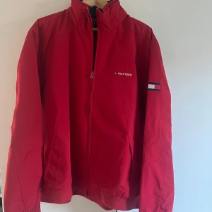 Röd jacka från Tommy Hilfiger - Säljer en snygg röd jacka från Tommy Hilfiger med dragkedja och logotyp på bröstet och kragen. Jackan har långa ärmar och en stilren design. Perfekt för att hålla stilen när det är kyligt ute.