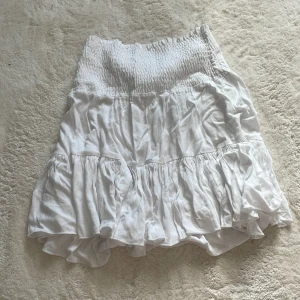 Vit volangkjol - Säljer en söt vit volangkjol med elastisk midja. Kjolen har flera lager av volanger som ger en luftig och lekfull känsla. Perfekt för varma dagar och kan enkelt matchas med olika toppar.