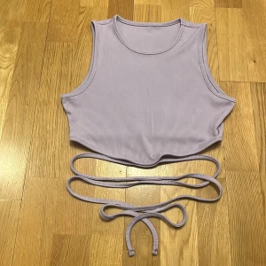 Ljuslila ribbad linnetopp - Säljer en snygg ljuslila ribbad linnetopp med knytband i midjan. Toppen är ärmlös och har en croppad passform, perfekt för sommardagar. Den mjuka ribbade texturen ger en stilren look.