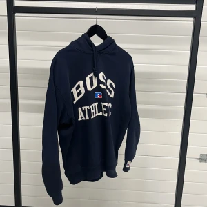 Mörkblå hoodie från Hugo Boss x Russell Athletic - Säljer en mörkblå hoodie från Hugo Boss i samarbete med Russell Athletic. Tröjan har en stor logotyp med texten 'BOSS ATHLETIC' på framsidan och en klassisk huva med dragsko. Perfekt för en sportig look! 