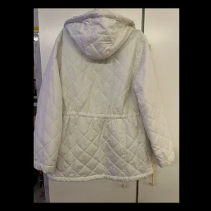 Vit quiltad jacka med huva - Säljer en stilren vit quiltad jacka med huva. Jackan har långa ärmar och praktiska fickor framtill. Den stängs med dragkedja och knappar, och har justerbar dragsko i midjan för bättre passform. Perfekt för kyligare dagar!