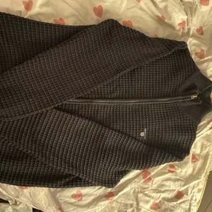 Svart ribbad tröja från GANT - Säljer en stilren svart ribbad tröja från GANT med dragkedja. Perfekt för kyligare dagar med sin långärmade design. Den har en diskret logga på bröstet som ger en exklusiv touch.