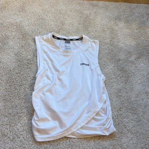 Vit ärmlös topp från Stronger - Säljer en vit ärmlös topp från Stronger i storlek XS. Toppen är tillverkad av 90% återvunnen polyester och 10% elastan, vilket ger en skön och stretchig passform. Perfekt för träning eller en aktiv vardag.