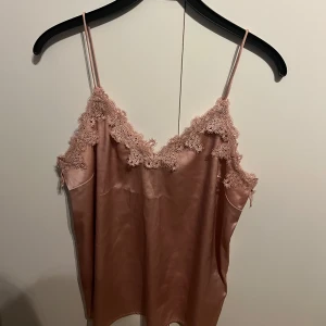 Rosa satinlinne från Bik Bok - Säljer ett elegant rosa satinlinne från Bik Bok med spetsdetaljer längs halsringningen. Linnet har tunna axelband och en mjuk, glansig yta. Perfekt för en stilren look.