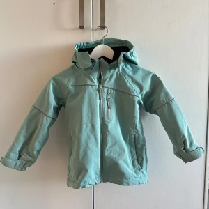 Grön vindjacka med huva barn  - Snygg grön vindjacka med huva och dragkedja framtill. Jackan har långa ärmar och praktiska fickor. Perfekt för blåsiga dagar.