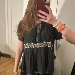 Odd Molly topp - Snygg svart blus med korta ärmar och dekorativ spetsdetalj i midjan från odd molly. Blusen har en knytning framtill med tofsar som ger en bohemisk känsla. Perfekt för en avslappnad stil.