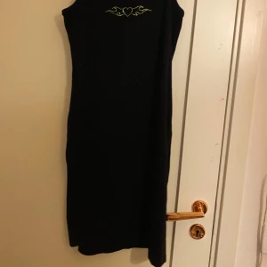 Svart ärmlös klänning från Lindex - Snygg svart ärmlös klänning från Lindex med ett dekorativt hjärtmönster i gult på bröstet. Perfekt för en stilren look.
