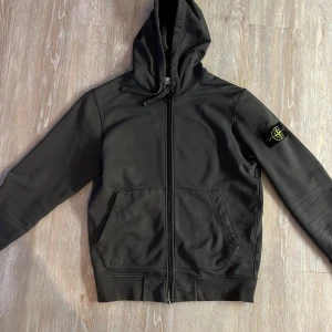 Svart hoodie från Stone Island - Snygg svart hoodie från Stone Island med dragkedja och huva. Tröjan har en ikonisk logotyp på ärmen och praktiska fickor framtill. Perfekt för en stilren och avslappnad look. Nypris 4000kr. 