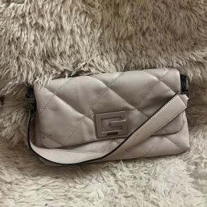 Beige quiltad axelväska från Guess - Snygg beige axelväska från Guess med quiltad design och ett stort G-emblem på framsidan. Väskan är perfekt för att bära dina nödvändigheter med stil.