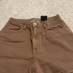 Bruna shorts från H&M - Säljer ett par snygga bruna shorts från H&M. Shortsen har en klassisk femficksdesign och är högmidjade med dragkedja och knapp framtill. Perfekta för en stilren look.