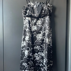 Svart och vit blommig klänning - Elegant svart klänning med vita blommor och smala axelband. Klänningen har en dekorativ rosett framtill och ett vackert blommönster som ger en stilren look.