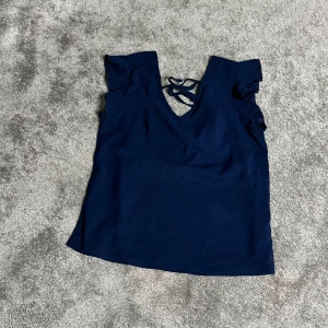 Mörkblå topp med volangdetaljer - Snygg mörkblå topp med volangdetaljer på axlarna och en elegant snörning i ryggen. Perfekt för en stilren look.