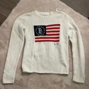 Vit stickad tröja från Bondelid - Säljer en vit stickad tröja från Bondelid med ett amerikanskt flaggmotiv på framsidan. Tröjan har långa ärmar och en rund halsringning. Perfekt för en avslappnad stil med en touch av retro.