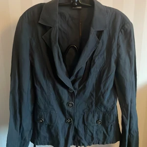 Marinblå kavaj från Gerry Weber Edition - Elegant marinblå kavaj från Gerry Weber Edition med klassisk krage och knappar framtill. Perfekt för en stilren look. Kavajen har långa ärmar och praktiska fickor.