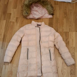 Ljusrosa dunjacka från Vinson Polo Club - Säljer en ljusrosa dunjacka från Vinson Polo Club med avtagbar huva och fuskpäls. Jackan har dragkedja och knappar framtill samt två fickor med dragkedja. Perfekt för kyliga dagar! Nypris 600 kr.