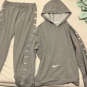 Grå hoodie och byxor från Nike - Säljer en grå hoodie och matchande byxor från Nike. Hoodien har en klassisk design med Nike-loggan på bröstet och längs ärmarna. Byxorna har elastisk midja och loggor längs benen. Perfekt för en avslappnad stil.