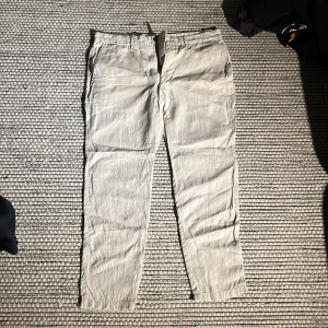 Beige linnebyxor från MNG - Snygga beige linnebyxor från MNG med en avslappnad passform. Perfekta för varma dagar med sitt lätta material. Byxorna har en klassisk design med dragkedja och knapp. Perfekt för en stilren look. Tyvärr har byxorna en fläck fram på höger smalben