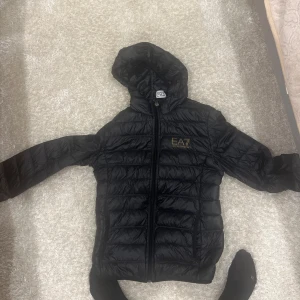 Svart quiltad jacka från EA7 - Snygg svart quiltad jacka från EA7 med huva och dragkedja. Perfekt för kyligare dagar med sin stilrena design och praktiska funktioner.
